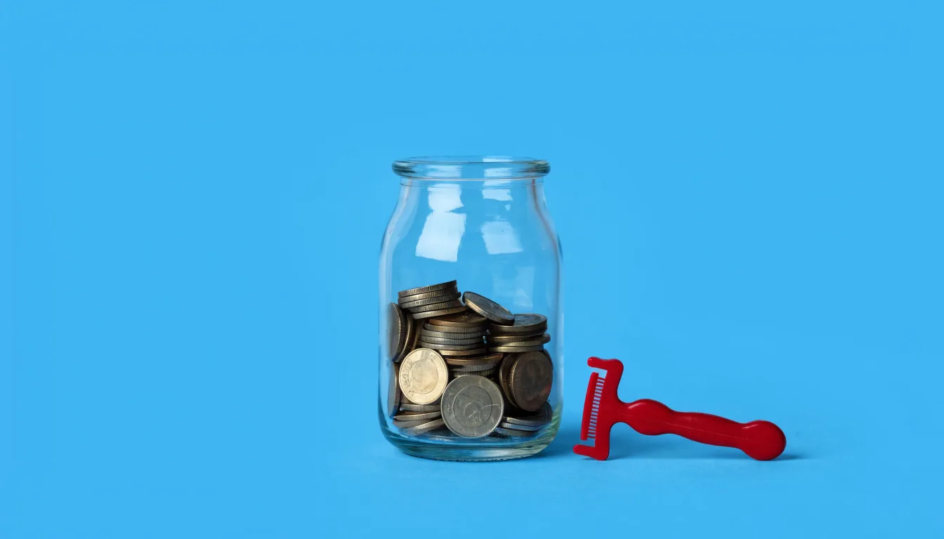 A small clear glass jar loosely filled with gold and silver coins, with a tiny red plastic toy razor resting visibly beside it, on a flat solid sky-blue background.