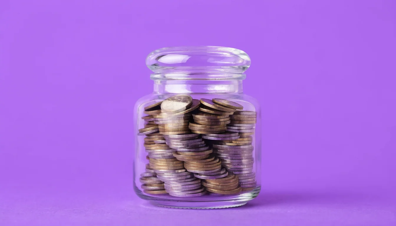 A clear glass jar filled with stacked gold and silver coins, centred on a flat solid purple background.