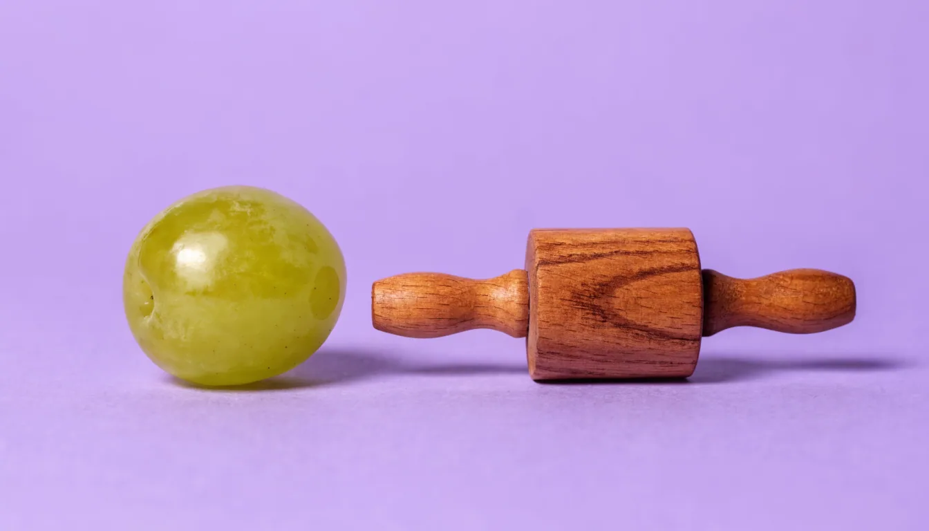 A plump whole green grape and a small wooden rolling pin placed side by side on a flat solid lavender background.