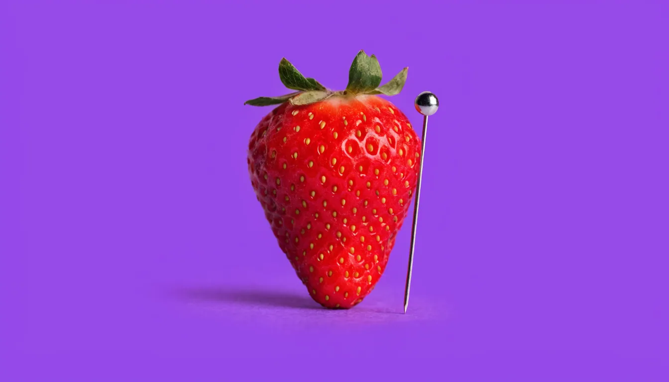 A whole fresh red strawberry with a single slim sewing pin resting lightly against its surface, on a solid purple background.